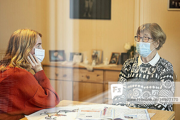 Enkelin und Mutter mit Gesichtsmaske im Gespräch  während sie mit sozialer Distanz zu Hause sitzen  während Covid-19