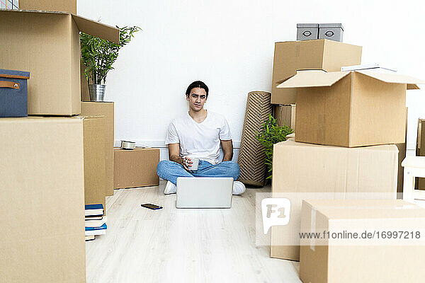 Junger Mann trinkt Kaffee und sitzt auf dem Boden  umgeben von Kisten in einer neuen Wohnung