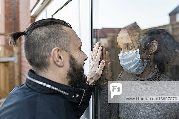 Mann sieht seine Freundin mit Gesichtsschutzmaske durch das Fensterglas während COVID-19