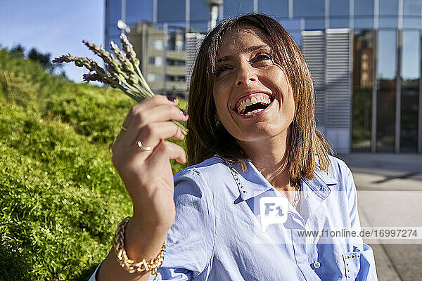 Weibliche Unternehmerin lacht  während sie an einem sonnigen Tag eine Lavendelpflanze an ein Bürogebäude hält