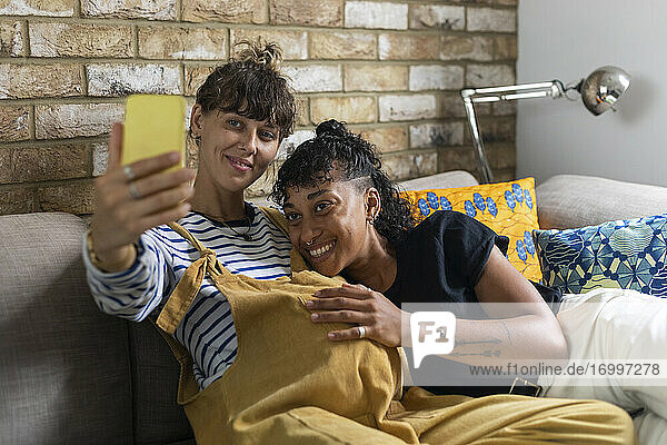 Schwangere Frau macht Selfie auf dem Handy mit ihrer Freundin  während sie zu Hause auf dem Sofa sitzt