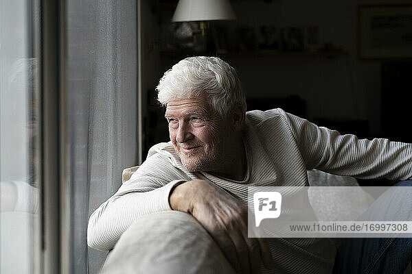 Lächelnder älterer Mann  der durch das Fenster schaut  während er zu Hause auf dem Sofa sitzt