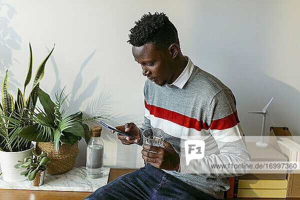 Männlicher Freiberufler  der mit seinem Smartphone SMS schreibt  während er im Büro zu Hause ein Trinkglas hält