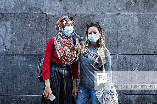 Frau mit Gesichtsmaske  während sie mit der Hand auf der Schulter eines muslimischen Freundes vor einer grauen Wand steht  während einer Pandemie