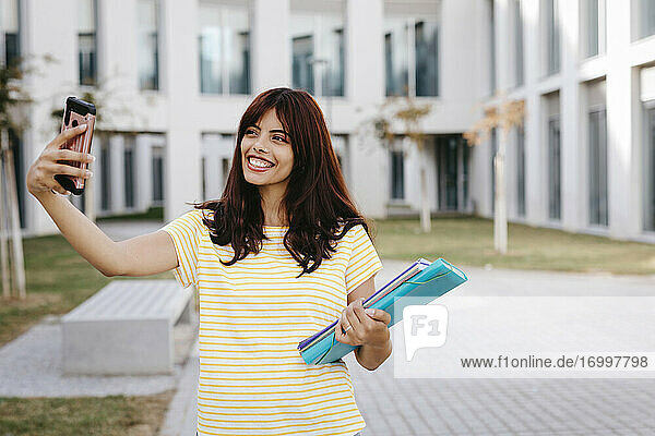 Smiling female student taking selfie with books at university campus