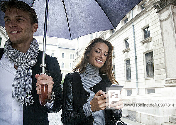 Lächelnde Geschäftsfrau  die unter einem Regenschirm mit einem männlichen Kollegen in der Stadt während der Regenzeit Textnachrichten auf einem Smartphone verschickt