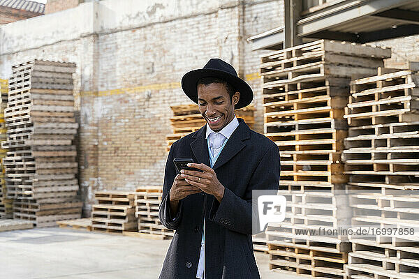 Junger Mann mit Hut  der ein Mobiltelefon benutzt  während er gegen Holzpaletten steht