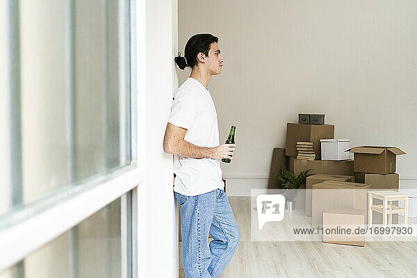 Nachdenklicher junger Mann  der eine Bierflasche hält  während er an einer Säule in einem Haus steht
