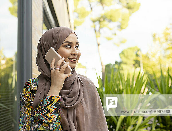 Junge Frau mit Hidschab  die im Freien steht und mit einem Mobiltelefon spricht
