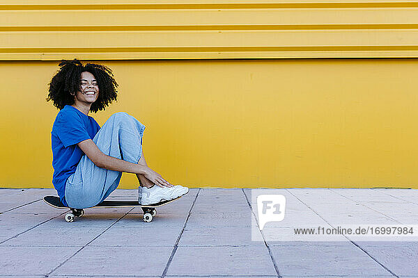 Lächelnde junge Frau sitzt auf einem Skateboard vor einer gelben Wand