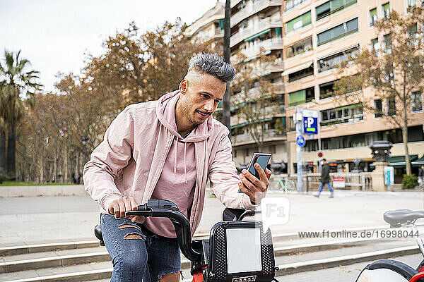 Stilvoller erwachsener Mann  der in der Stadt ein Fahrrad mit Mobiltelefon mietet