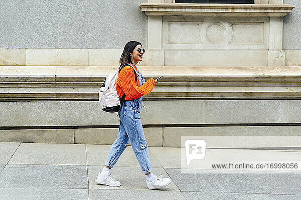Lächelnde Frau mit Rucksack  die ein Mobiltelefon benutzt  während sie auf einem Fußweg geht