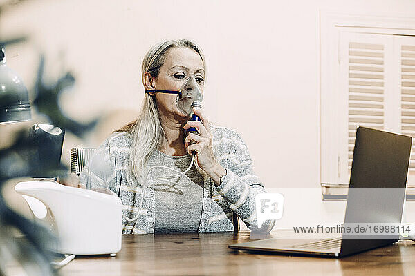 Ältere Frau  die eine Online-Konsultation in Anspruch nimmt  während sie zu Hause durch einen Vernebler inhaliert