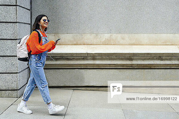 Lächelnde Frau mit Rucksack  die ein Mobiltelefon benutzt  während sie gegen eine Wand läuft