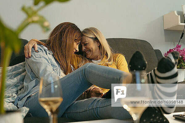 Smiling mother and daughter with arm around touching foreheads on sofa