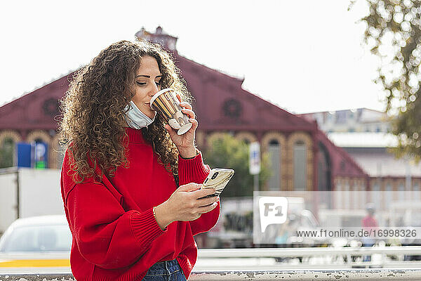Junge Frau trinkt Kaffee und benutzt ein Mobiltelefon in der Stadt während COVID-19