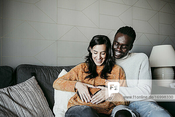 Glücklicher Mann und schwangere Frau bilden Herzform mit Händen auf dem Bauch  während sie auf dem Sofa zu Hause sitzen