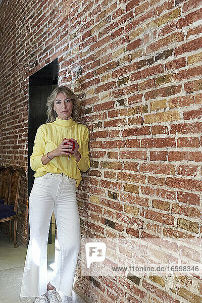 Mature woman with tea cup leaning on brick wall in kitchen