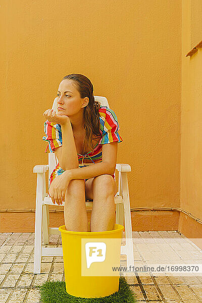 Porträt einer ernsten Frau  die sich auf einem Plastikstuhl ausruht und ihre Beine in einem Eimer Wasser kühlt