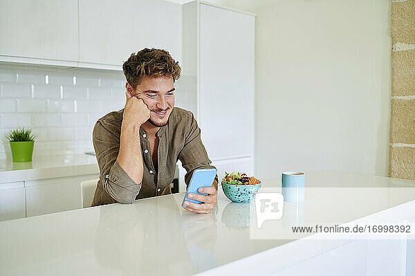 Lächelnder Mann  der sein Smartphone benutzt  während er sich zu Hause auf die Küchentheke stützt