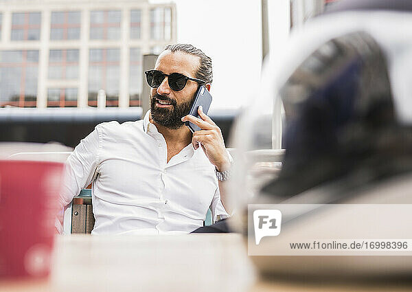 Porträt eines bärtigen Geschäftsmannes mit Sonnenbrille  der im Freien mit einem Smartphone telefoniert
