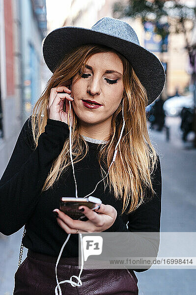 Blond woman wearing hat listening music through mobile phone while standing on street