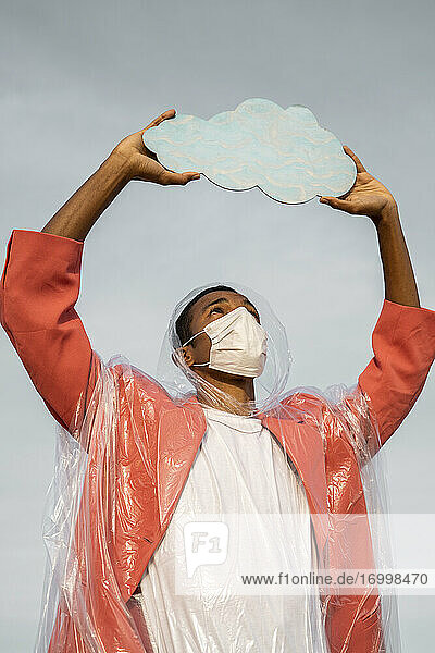 Man wearing protective face mask and raincoat holding cloud cut out while standing against sky