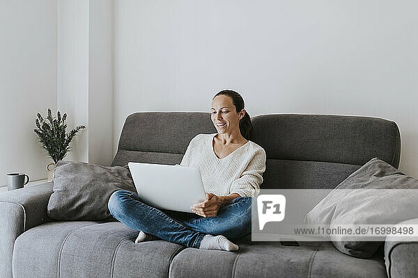 Lächelnde Frau  die vom Laptop aus einen Videogespräch führt  während sie zu Hause auf dem Sofa sitzt