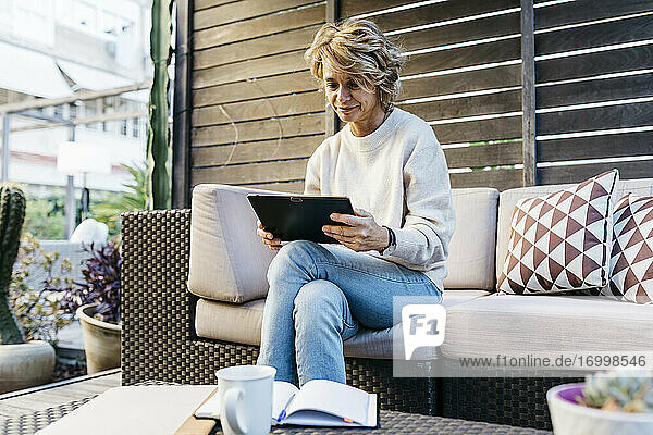 Frau  die ein digitales Tablet benutzt  während sie auf einem Sofa auf der Terrasse eines Gebäudes sitzt