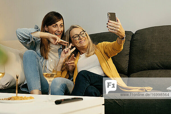 Smiling mother and daughter making peace sign while taking selfie through smart phone at home