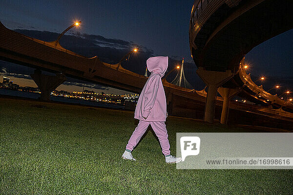 Frau in rosa Trainingsanzug und Kapuze  die nachts auf einer Wiese unter einer Hochstraße spazieren geht