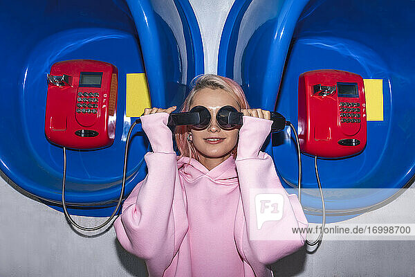 Lächelnde junge Frau mit rosa Haaren und rosa Kapuzenshirt steht an einer Telefonzelle und hält sich einen Hörer vor die Augen