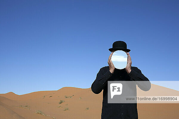 Marokko  Merzouga  Erg Chebbi  Mann mit Melone  der sich in einer Wüstendüne einen Spiegel vor das Gesicht hält