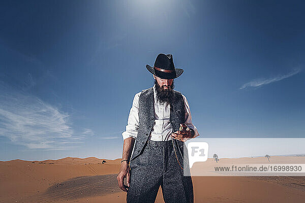 Man with a beard and hat in the dunes of the desert of Morocco checking the time