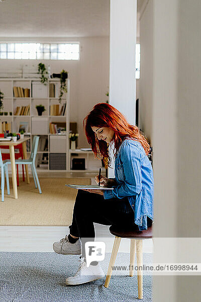 Junge Geschäftsfrau  die im Büro auf einem Hocker sitzend an einem digitalen Tablet arbeitet