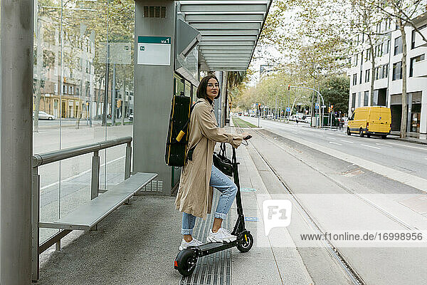 Frau mit Tretroller und Instrumentenkoffer an einer Straßenbahnhaltestelle in der Stadt