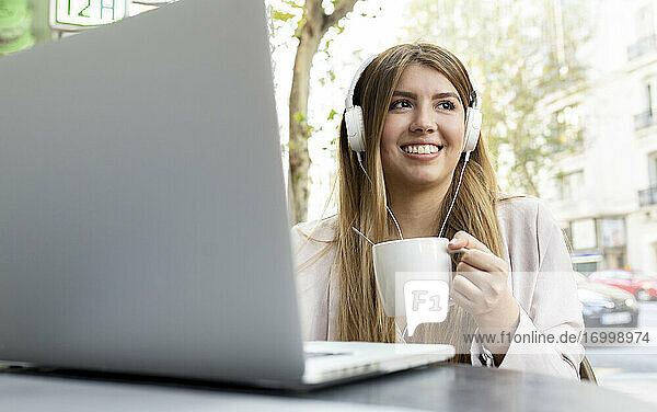 Frau mit Kopfhörern trinkt Kaffee  während sie mit einem Laptop in einem Straßencafé sitzt