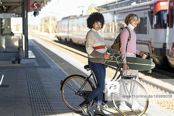Lächelnde männliche und weibliche Freunde  die mit Fahrrad und Longboard am Bahnhof warten  schauen weg