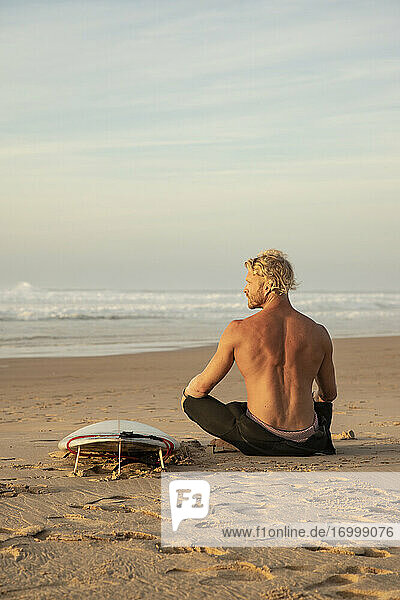 Shirtless surfer sitting by surfboard on sand while looking at sea against sky
