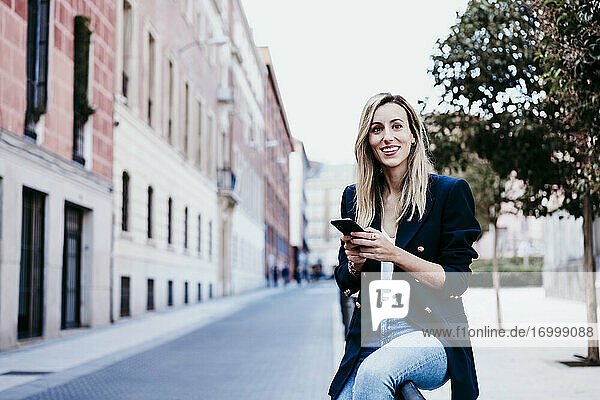 Lächelnde modische Frau  die wegschaut  während sie ein Mobiltelefon in der Stadt hält