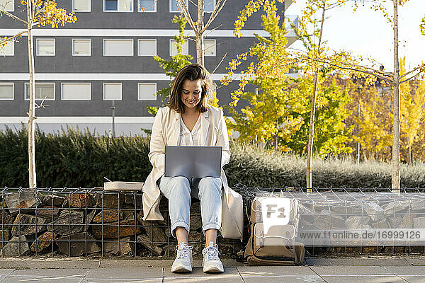Lächelnde Geschäftsfrau  die einen Laptop benutzt und auf einer Stützmauer in der Stadt sitzt