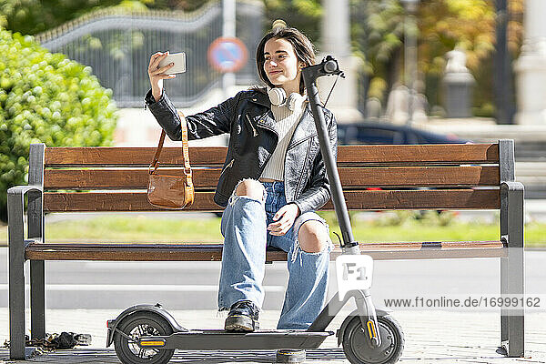 Smiling woman taking selfie through smart phone while sitting on bench by electric push scooter
