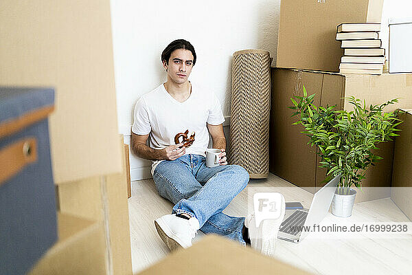 Junger Mann isst Churro mit Kaffee und sitzt in seiner neuen Wohnung