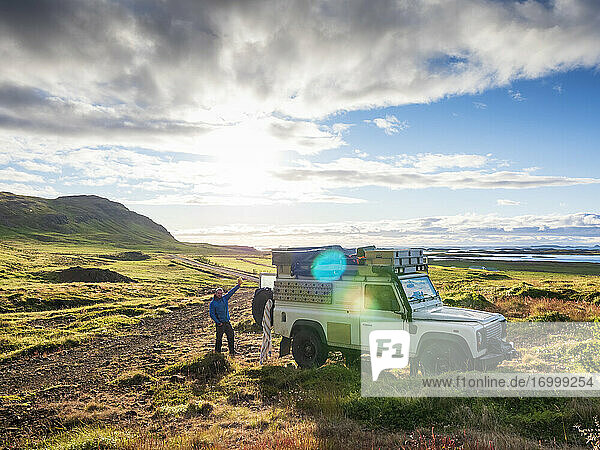 Sonnenuntergang über einem einsamen Reisenden  der hinter einem Geländewagen steht und der Kamera zuwinkt  Island