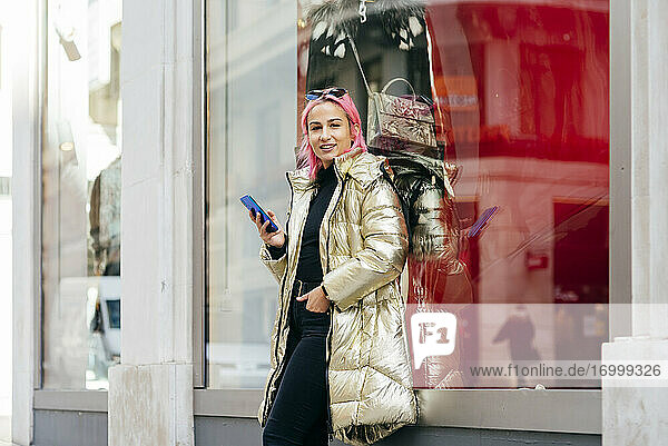 Modische Frau  die ein Mobiltelefon benutzt  während sie mit den Händen in den Taschen am Fenster steht