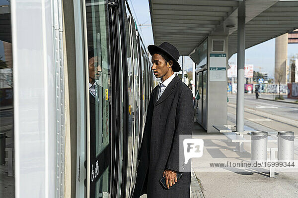 Modischer junger Mann  der an einem sonnigen Tag an der Straßenbahntür am Bahnhof steht