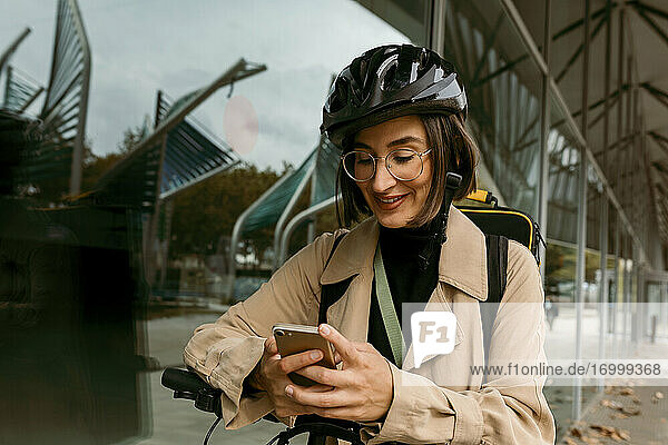 Lächelnde Frau mit elektrischem Tretroller  die ein Mobiltelefon benutzt  während sie an einer Glaswand steht