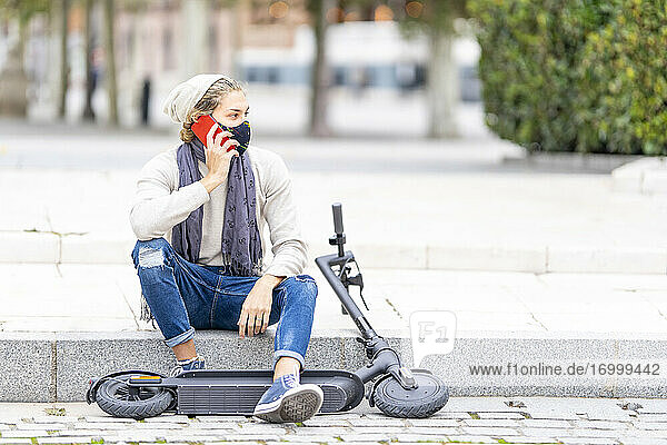 Junger Mann mit Gesichtsmaske  der mit einem Mobiltelefon spricht  während er auf einem elektrischen Roller in der Stadt sitzt