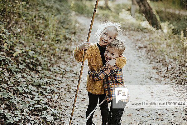 Sister holding stick while embracing brother standing in forest