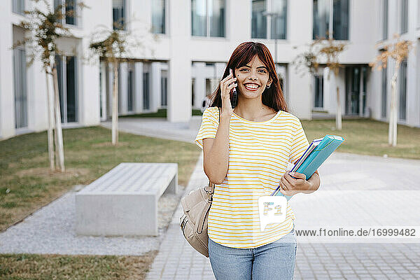 Lächelnde hispanische Studentin  die mit Büchern und einem Ordner steht und mit einem Mobiltelefon spricht